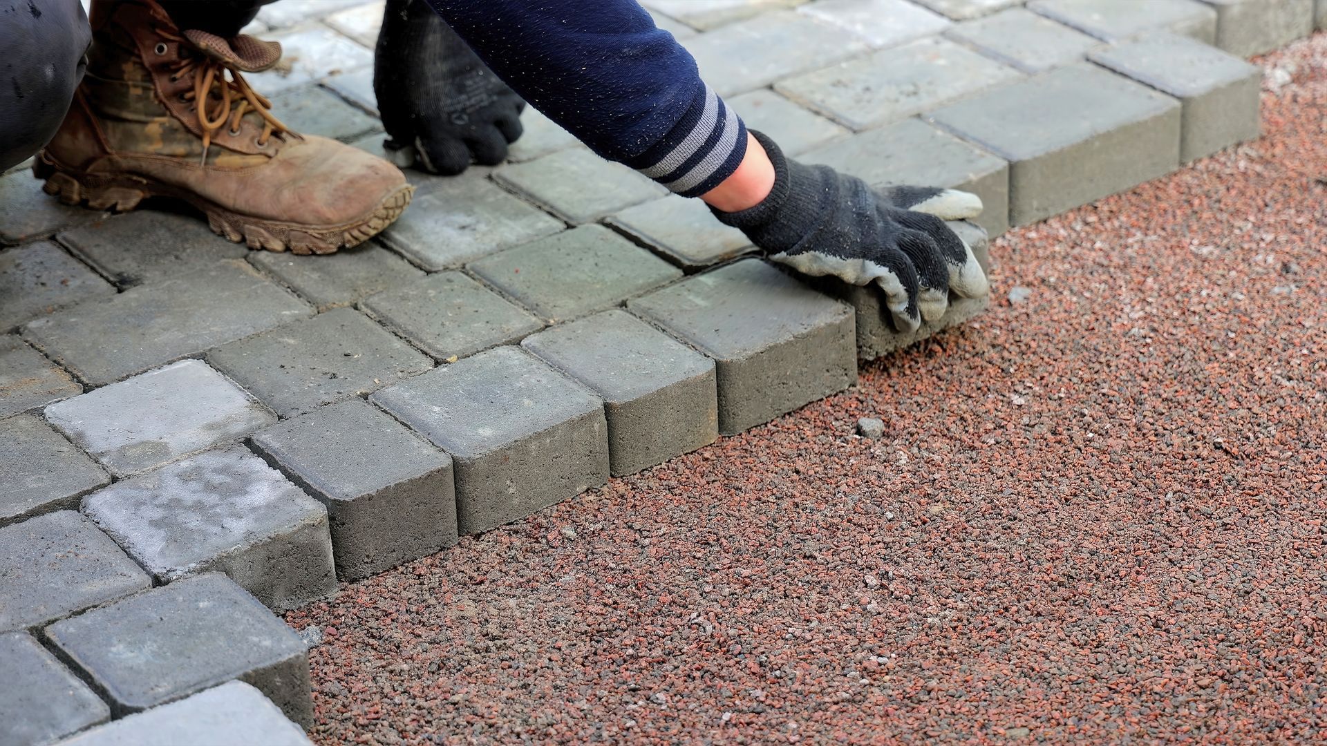 Person in work gloves laying gray paving stones on a red gravel base.