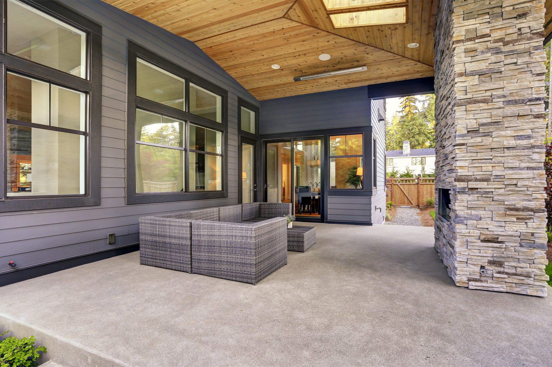 Covered patio with gray wicker furniture, gray siding, and a stone column fireplace.