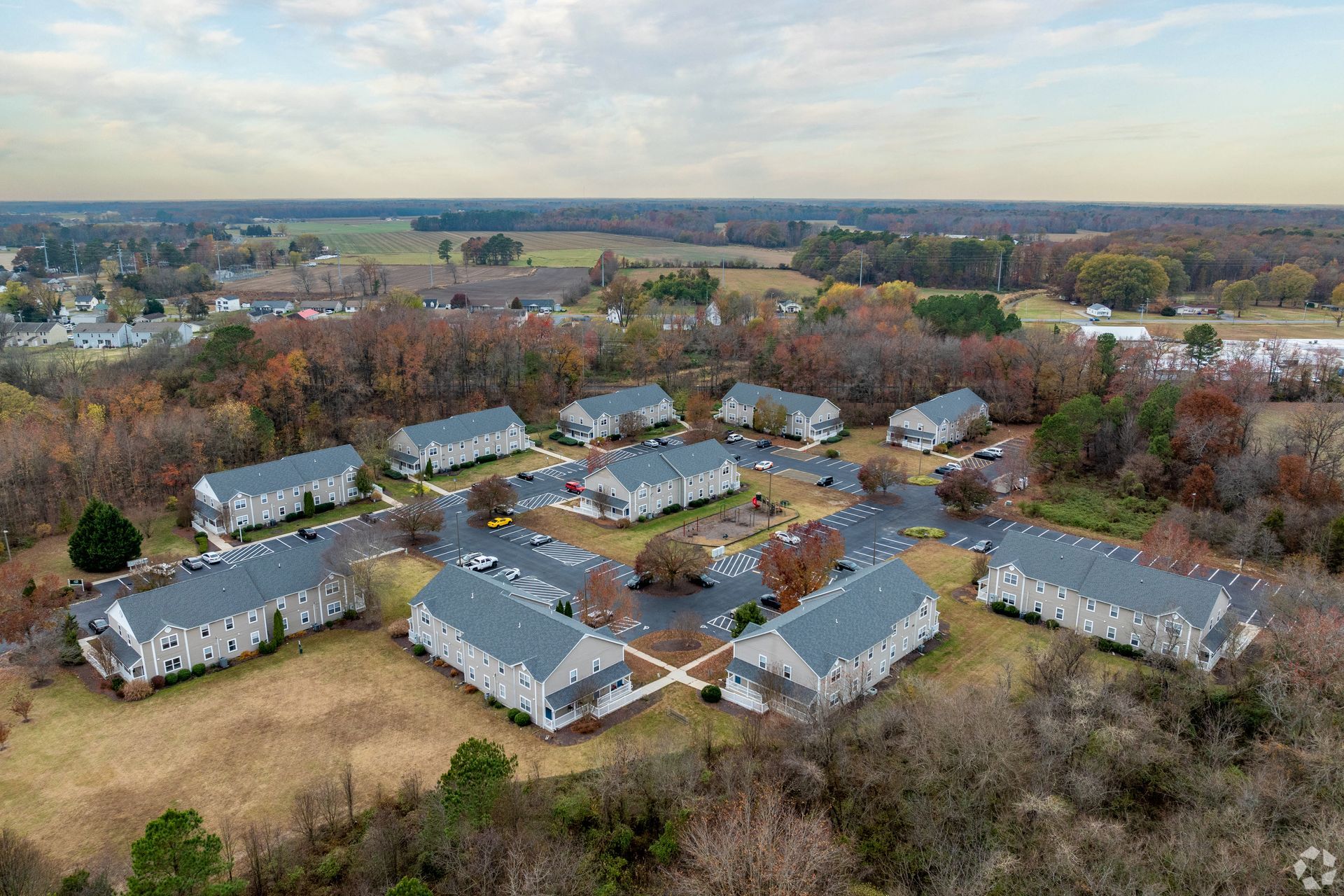 Aerial view of a circular housing development with gray buildings and a central parking area, surrounded by trees and fields.