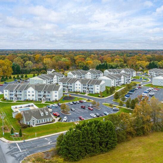 Aerial view of apartment complex with white buildings, parking, and a pool, surrounded by trees.
