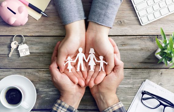 Hands holding paper family cutouts; table with keys, piggy bank, coffee, and office supplies.