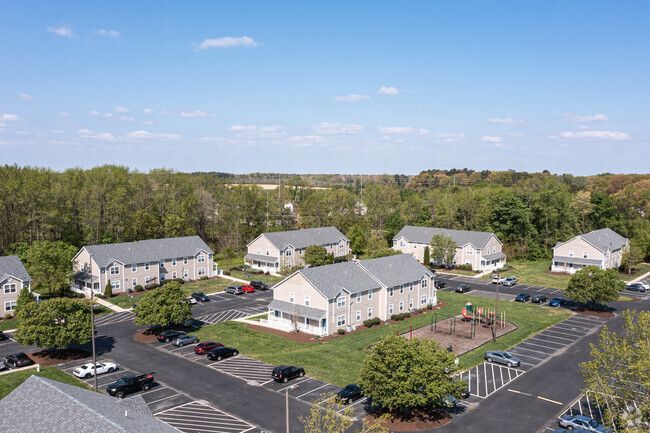 Aerial view of an apartment complex with gray roofs and light tan siding, surrounded by trees and parking lots on a sunny day.