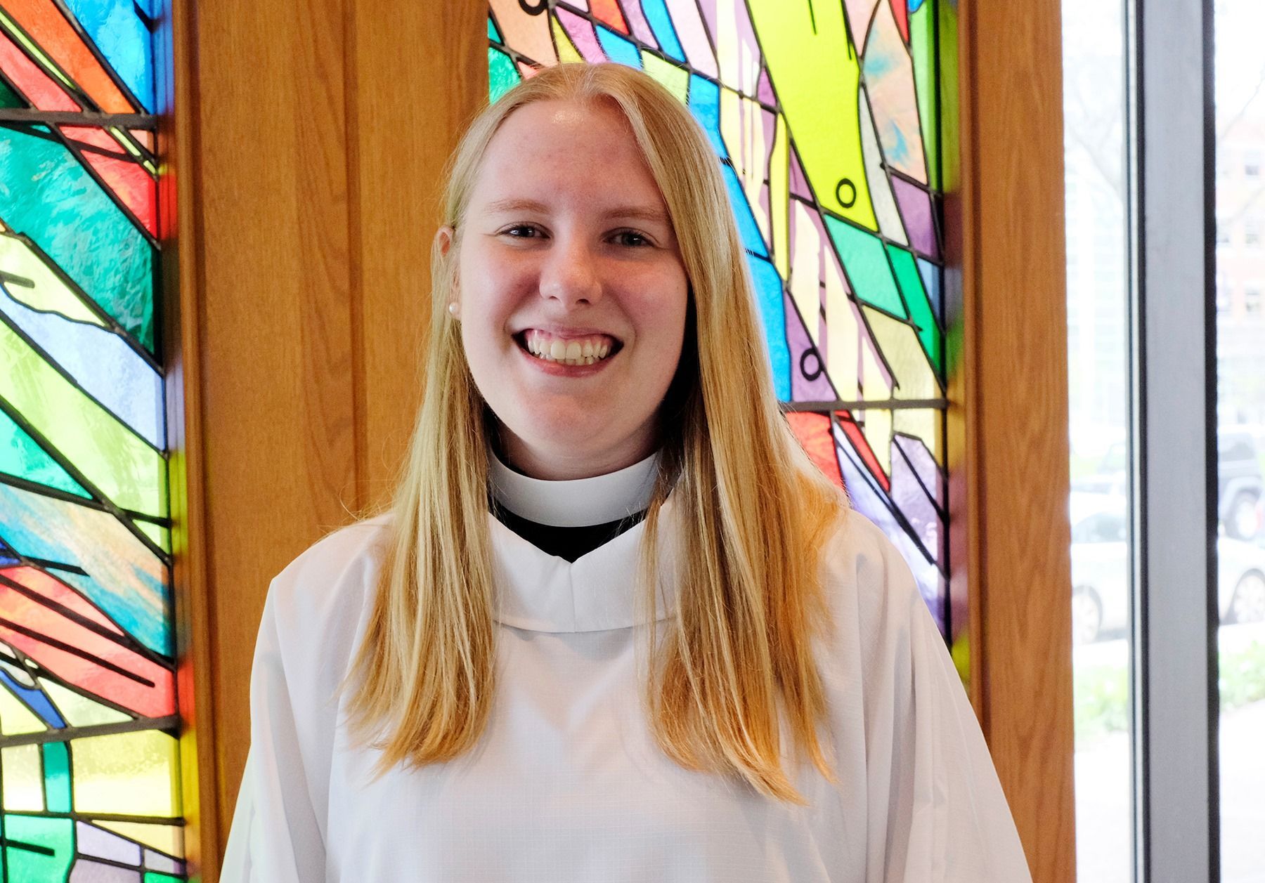 Smiling person in a white robe standing before colorful stained-glass windows.