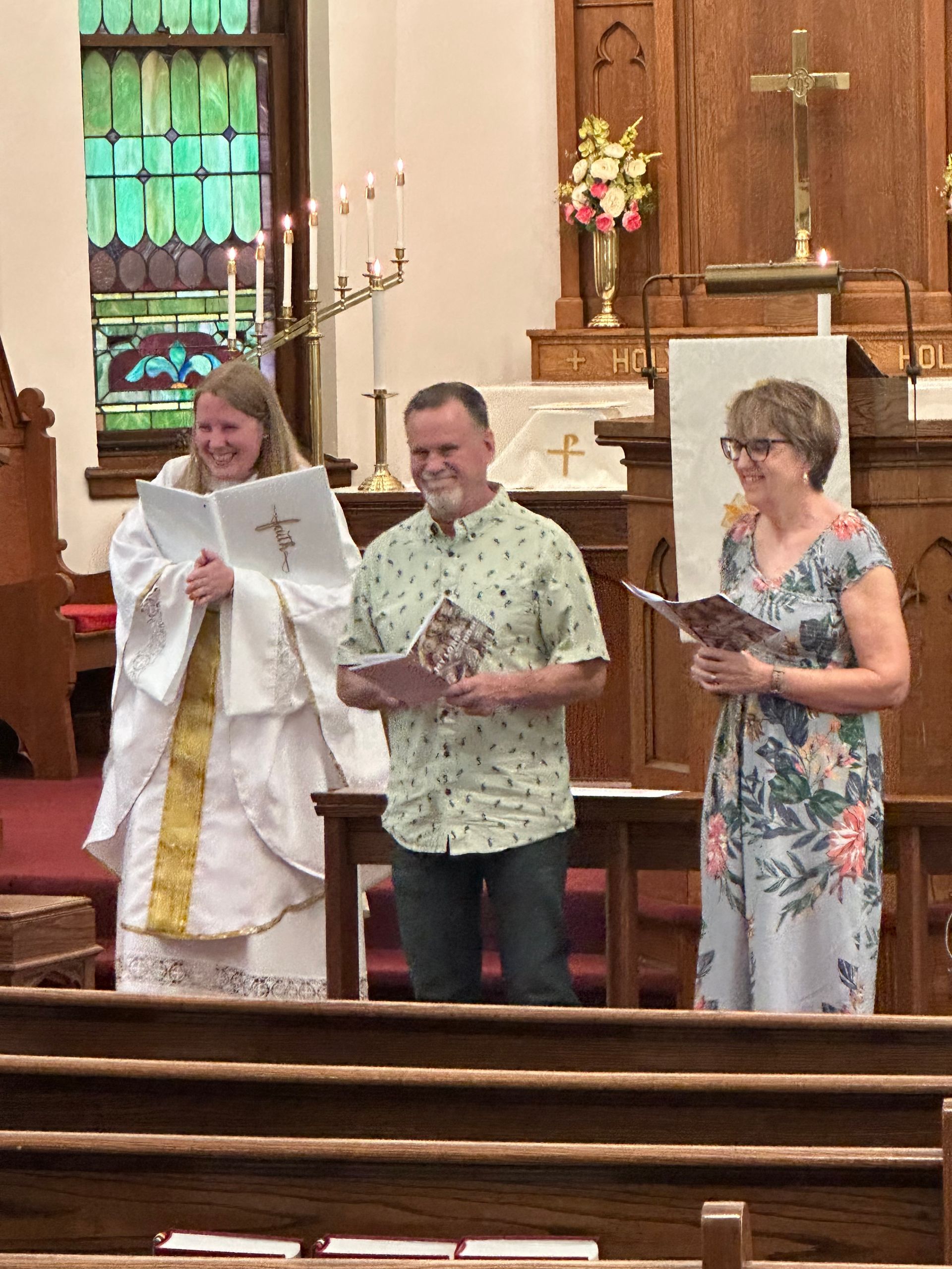 Three people stand at a church altar holding papers, one in a white robe, two in patterned clothes.