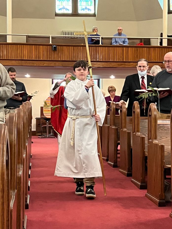 Child in white robe holding a candle, walking down a church aisle during a service.