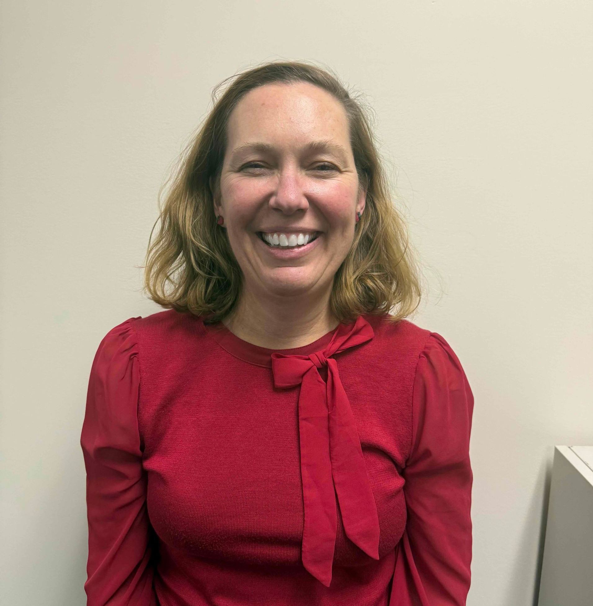 Smiling person in a red blouse standing against a beige wall.