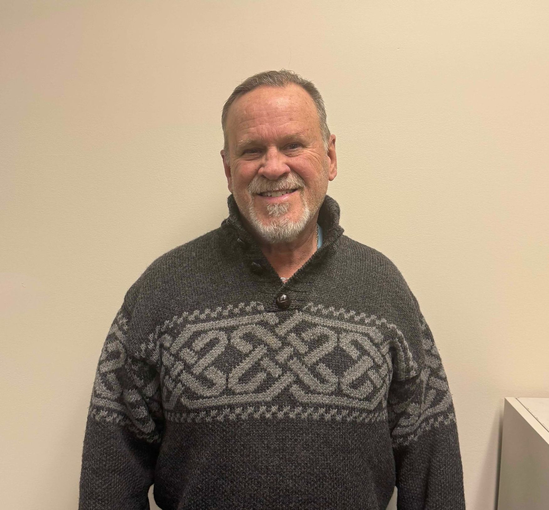 Smiling man in a gray patterned sweater standing against a plain beige wall
