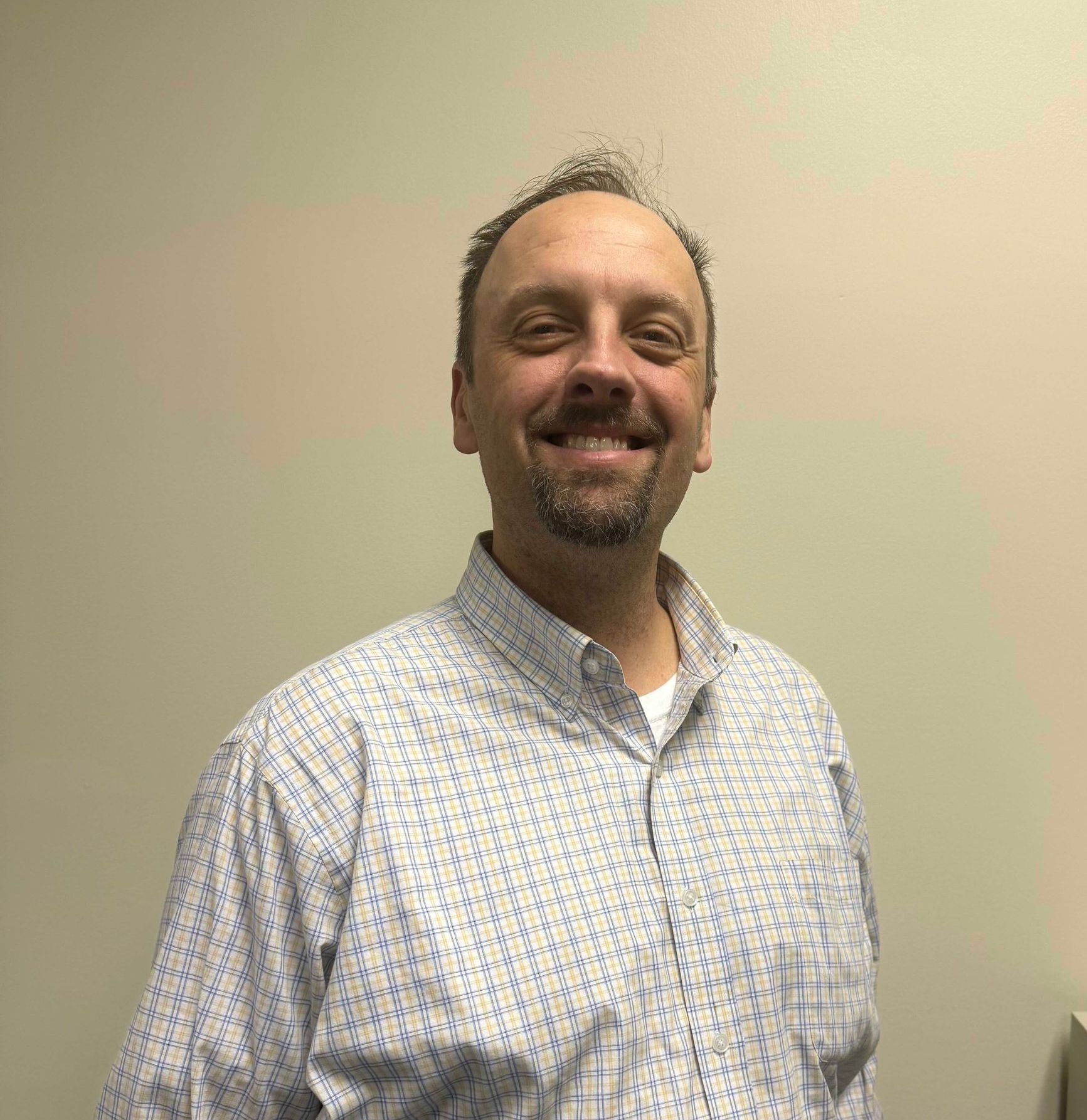 Smiling man in a white patterned shirt standing against a plain beige wall.
