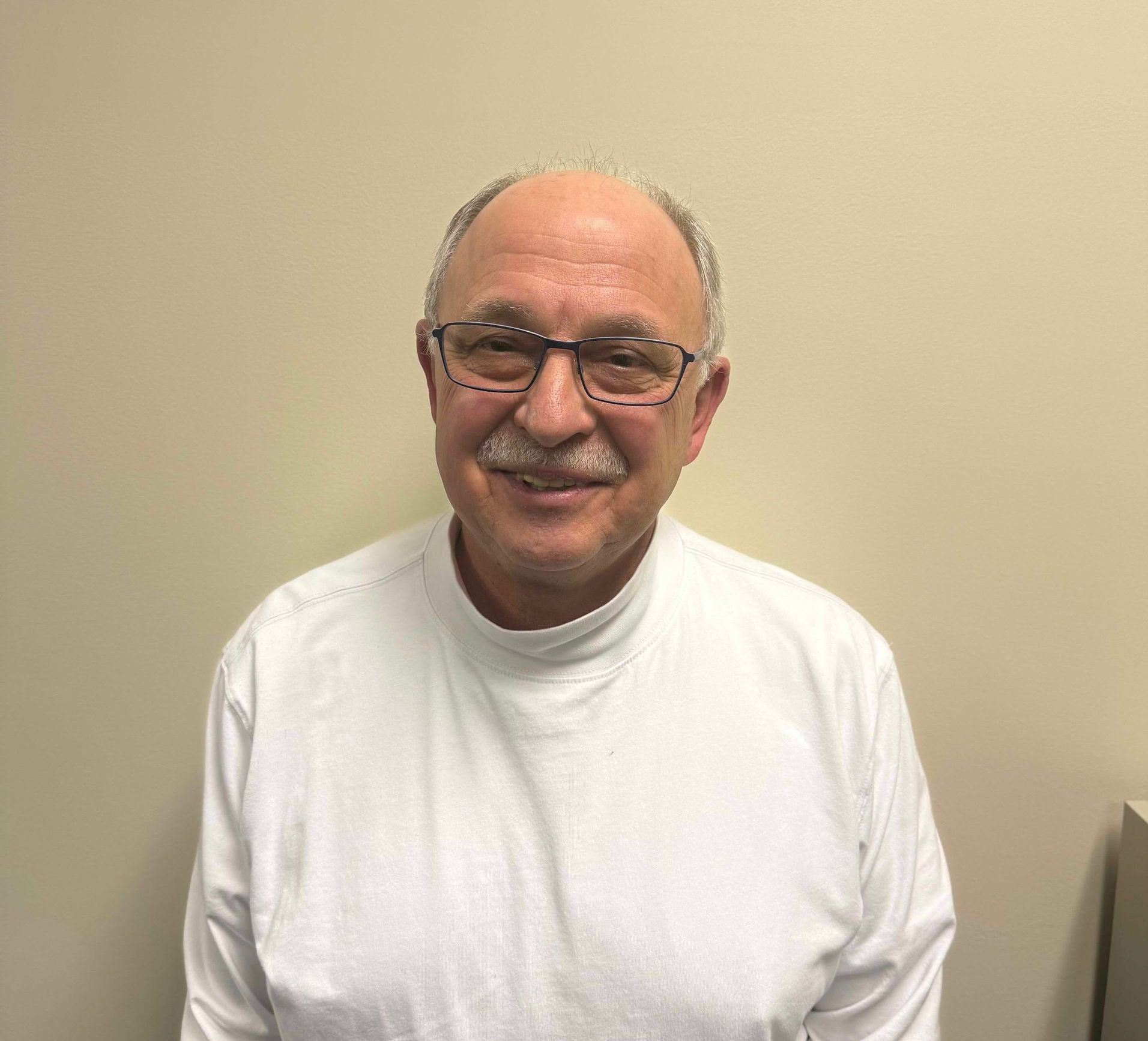 Smiling man wearing glasses and a white shirt against a plain beige wall.