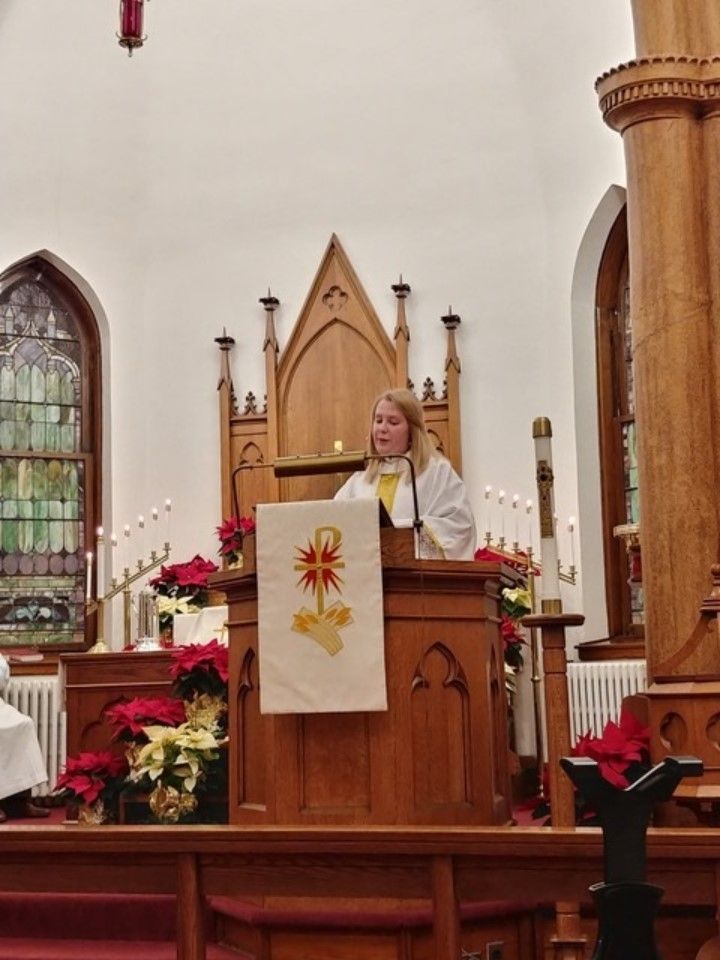 Woman speaking at a church pulpit with a lit Christmas altar and poinsettias behind her