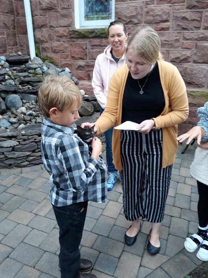 Woman handing a paper to a child outdoors by a stone wall, with another adult smiling in the background.
