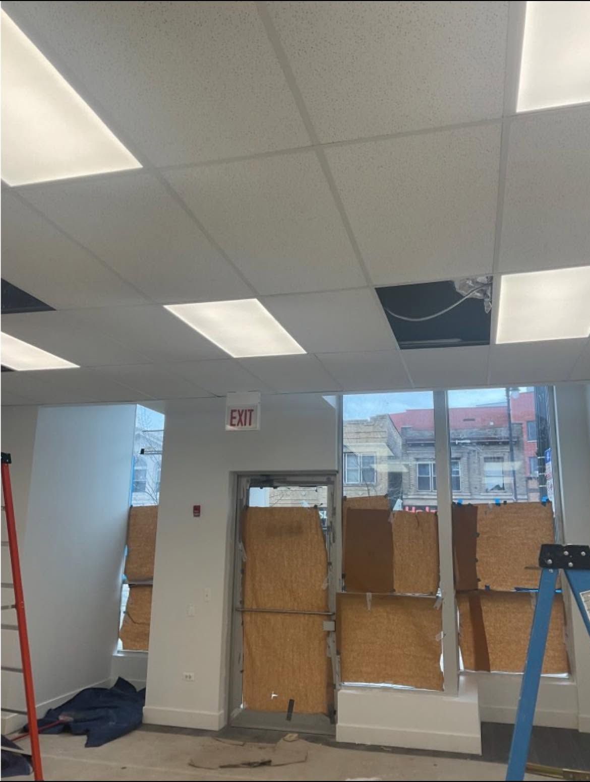 Office interior with damaged ceiling tiles, boarded windows, and blue ladder near an exit sign