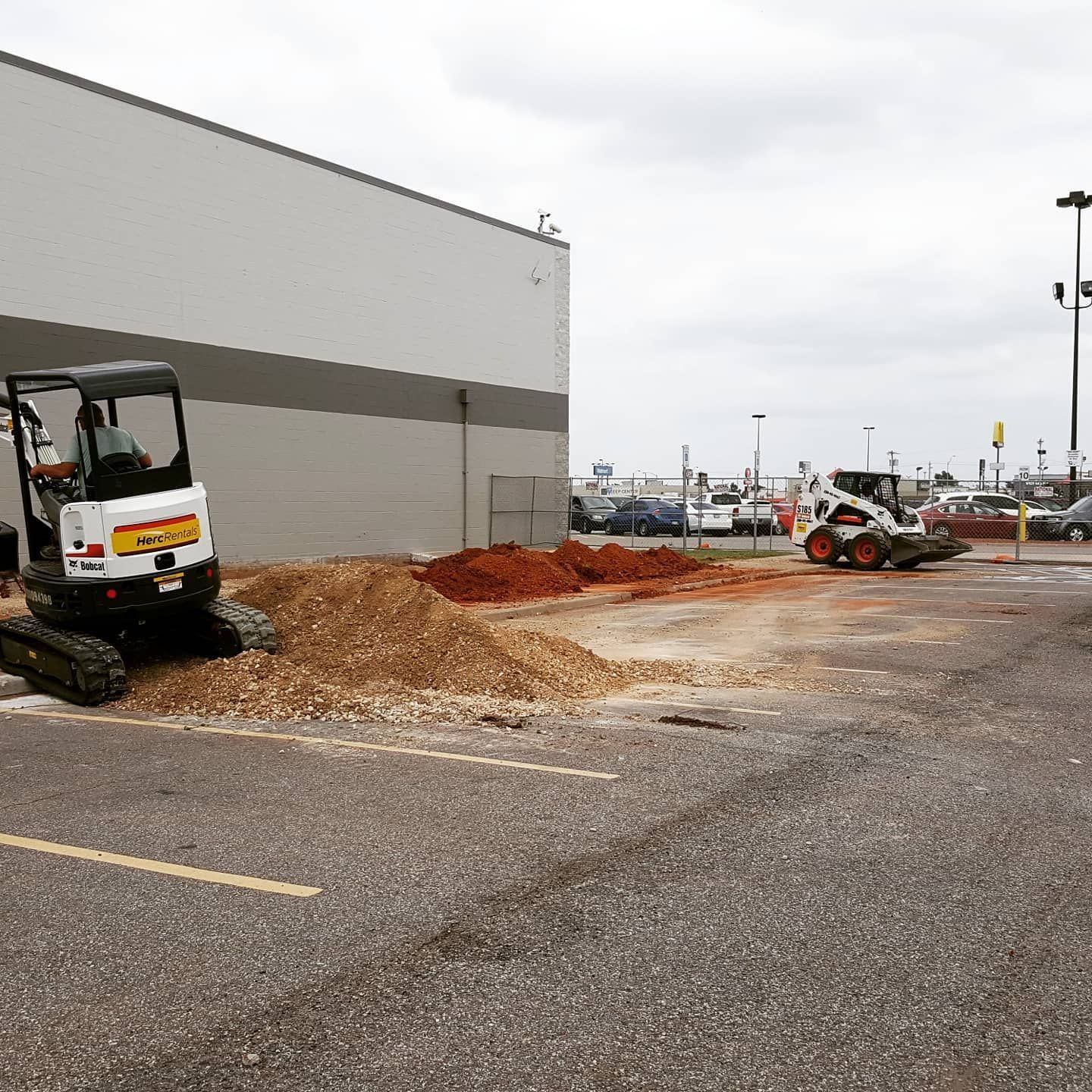 Construction site beside gray building with excavator and dirt piles in a parking lot