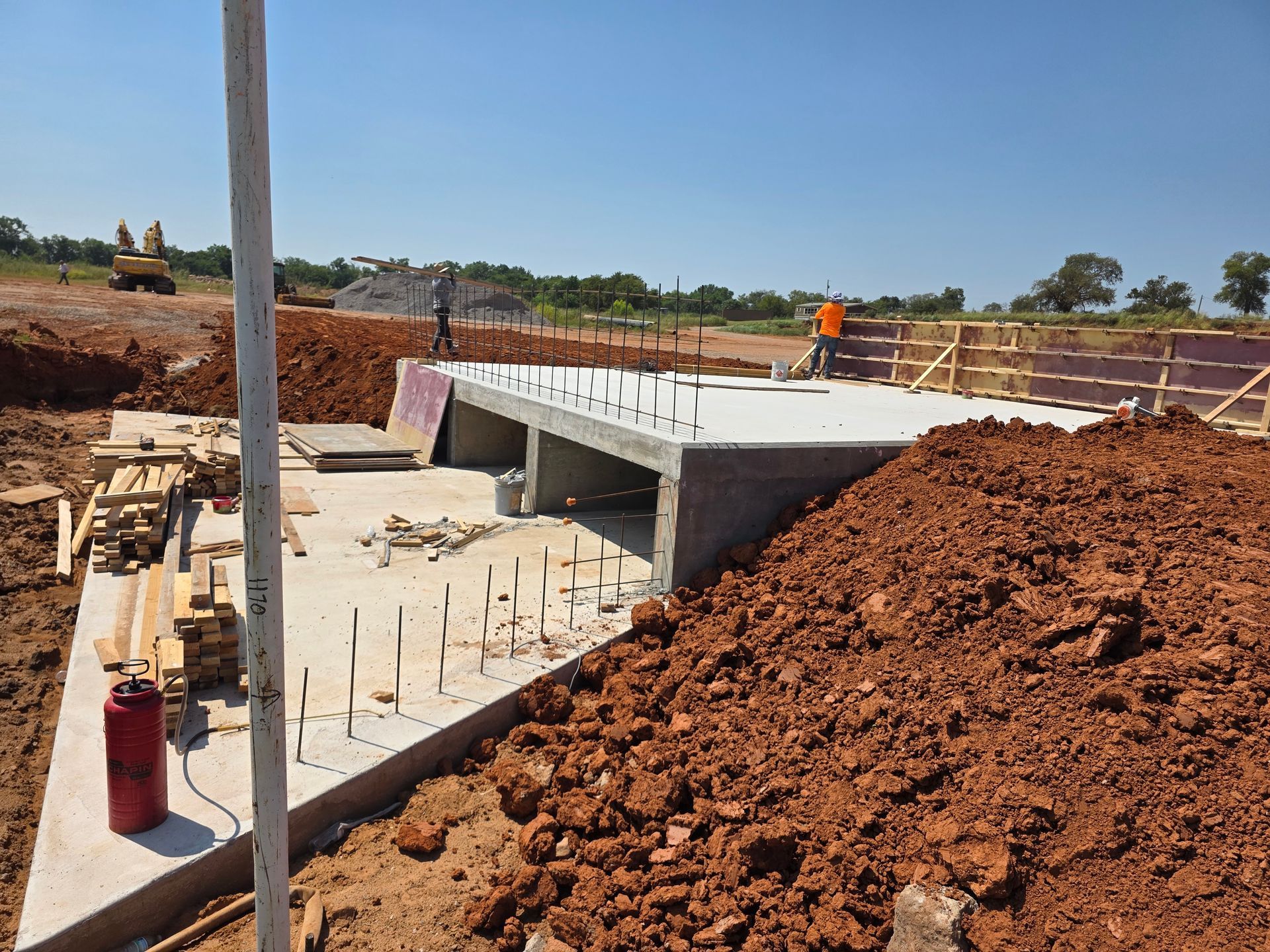 Construction site with concrete bridge or culvert, dirt embankments, and workers in orange safety gear