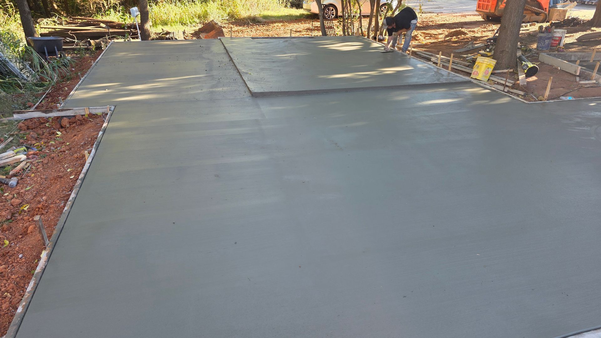 Freshly poured gray concrete driveway in a yard, with a worker and equipment in the background