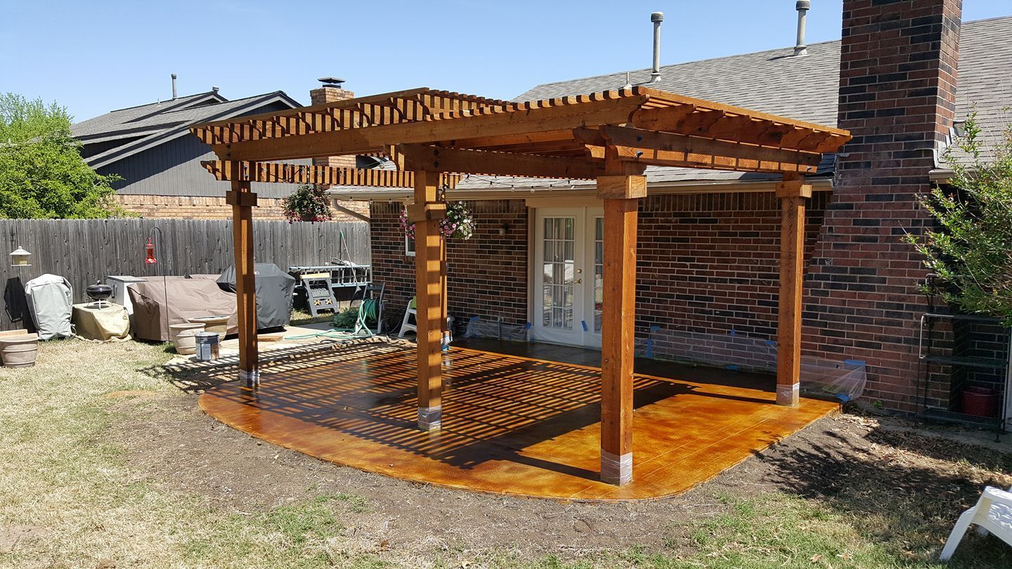 Wooden pergola patio beside a brick house, with a glossy deck and lattice-style roof under bright sun.