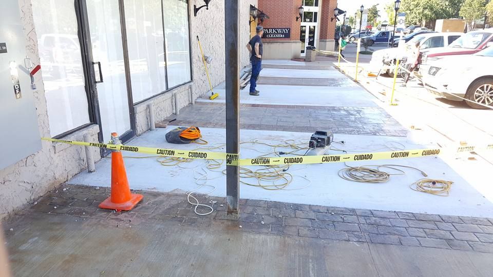 Caution tape blocks a cracked sidewalk beside a building, with orange cones and workers in the distance
