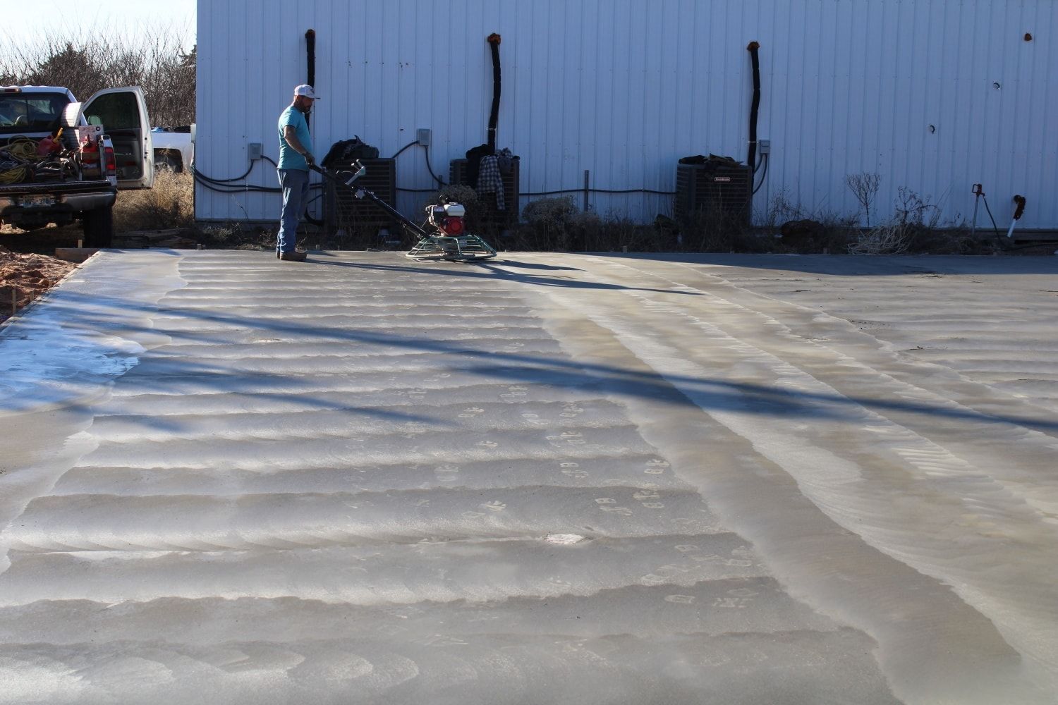 Worker on icy lot near a blue industrial building, with equipment and parked vehicles in the background