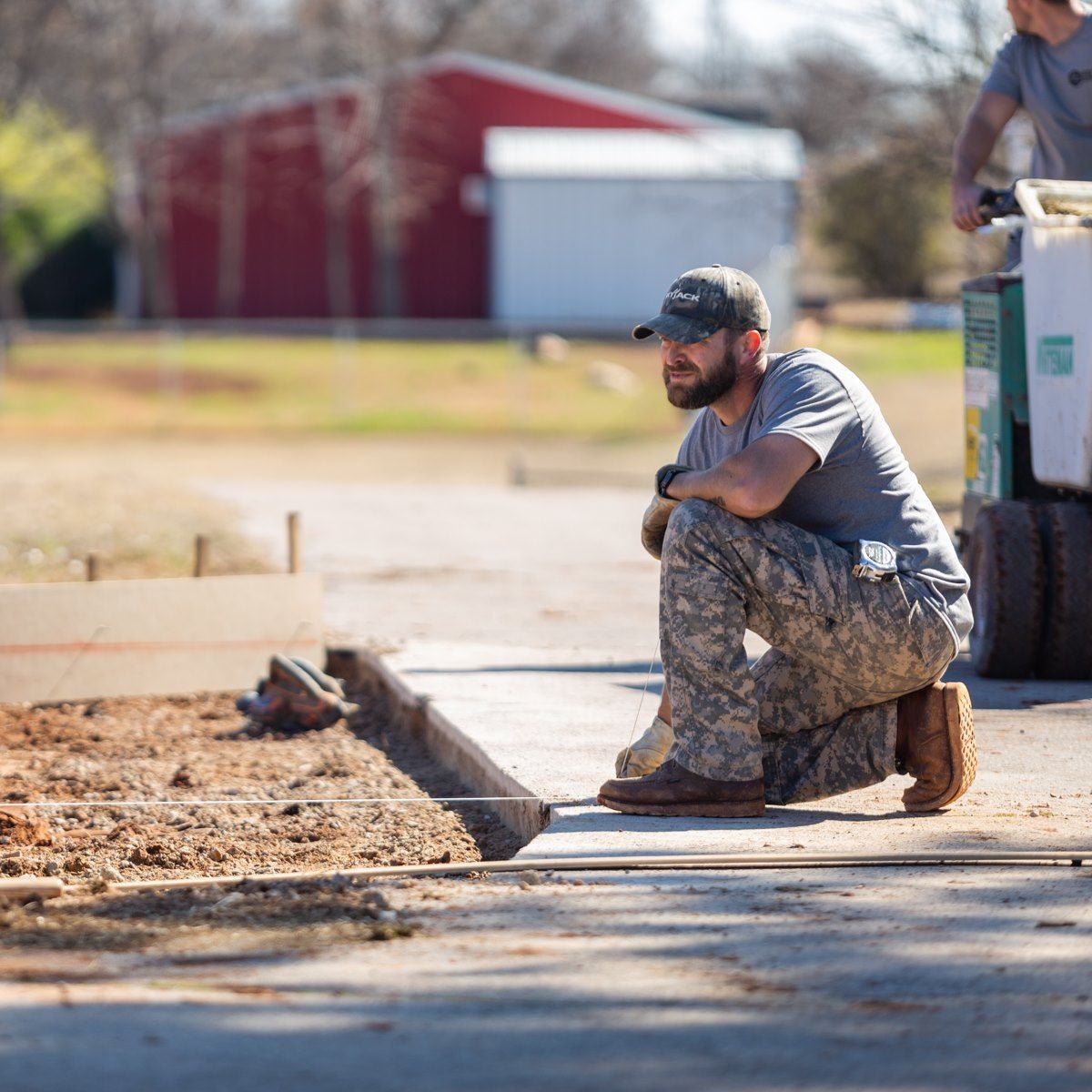Worker in camouflage kneels by a sidewalk near a dirt trench at a construction site.