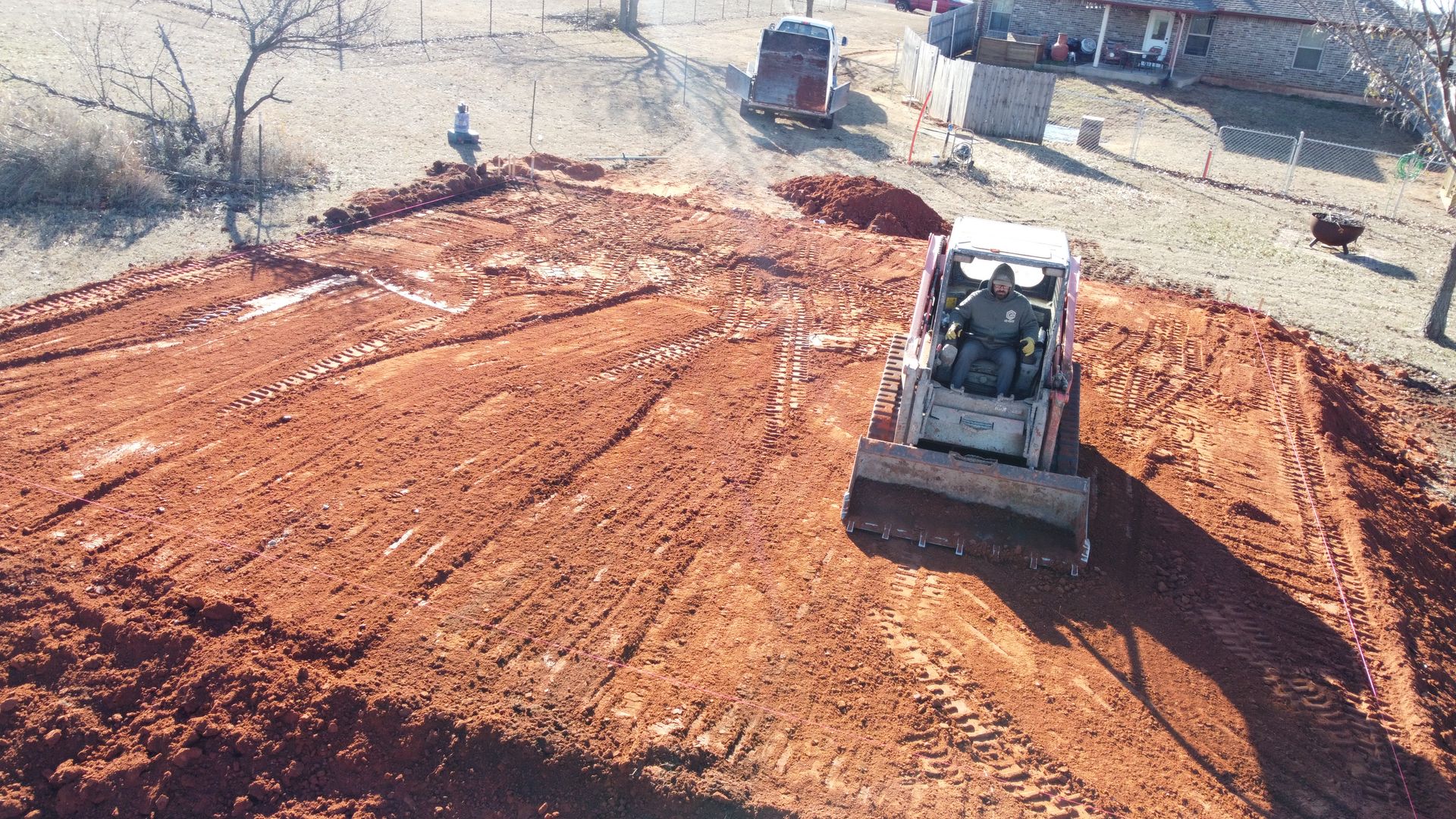 Construction site with a skid steer grading red dirt, with a truck and excavator in the background.
