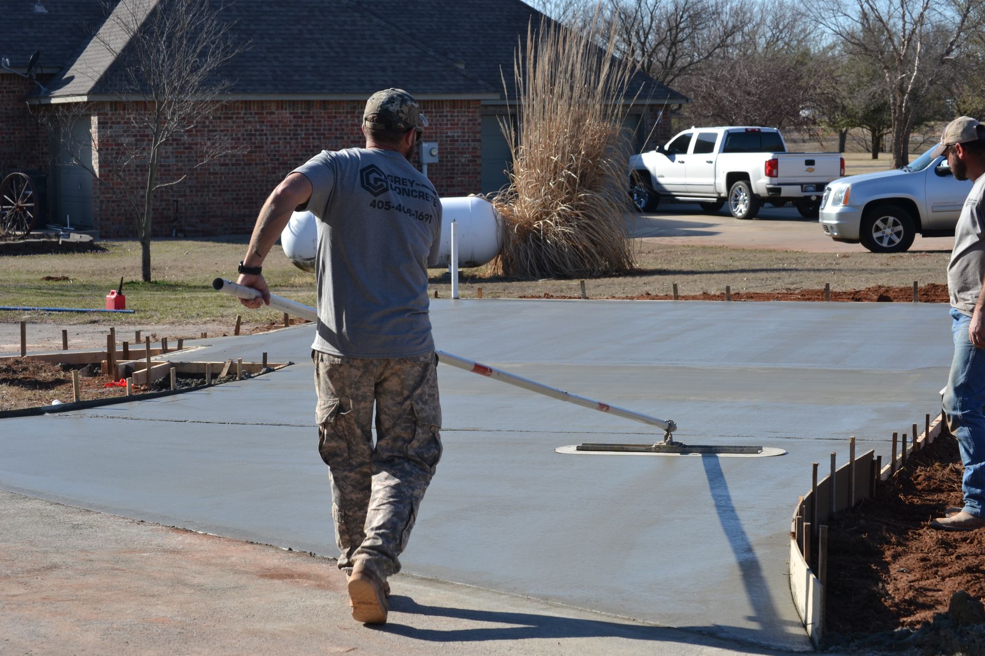 Worker smoothing fresh concrete driveway with a long-handled tool in a suburban yard