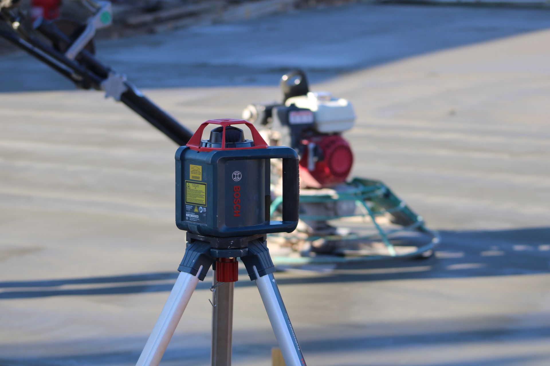 Surveying equipment on a tripod in a parking lot, with a red machine and cables in the background