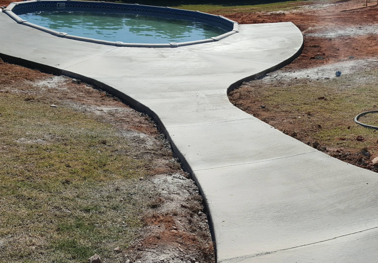 Curving concrete walkway leading to a small blue pool amid brown grass and mulch