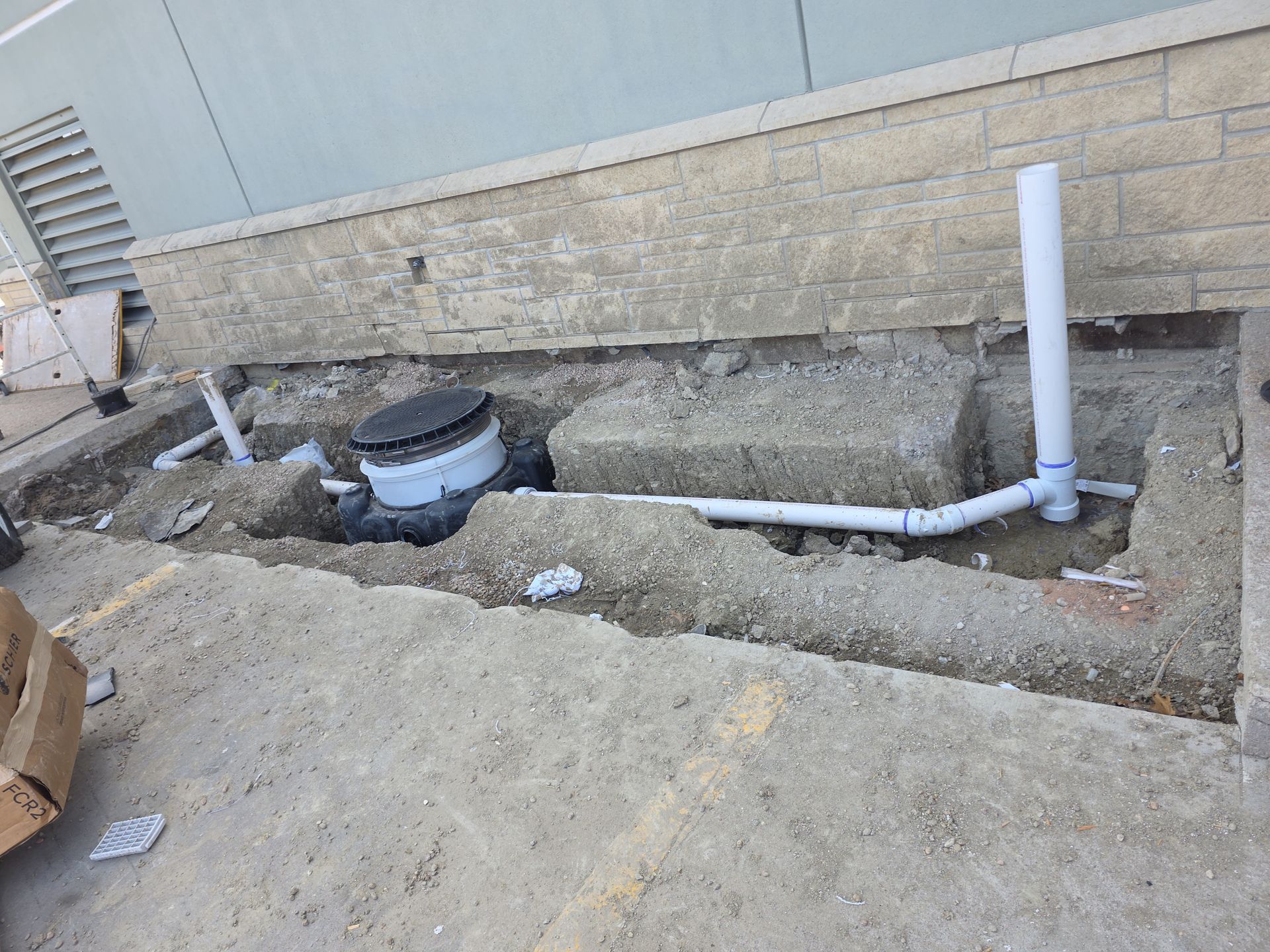 Uneven gravel trench beside a building wall with white PVC pipes and utility equipment.