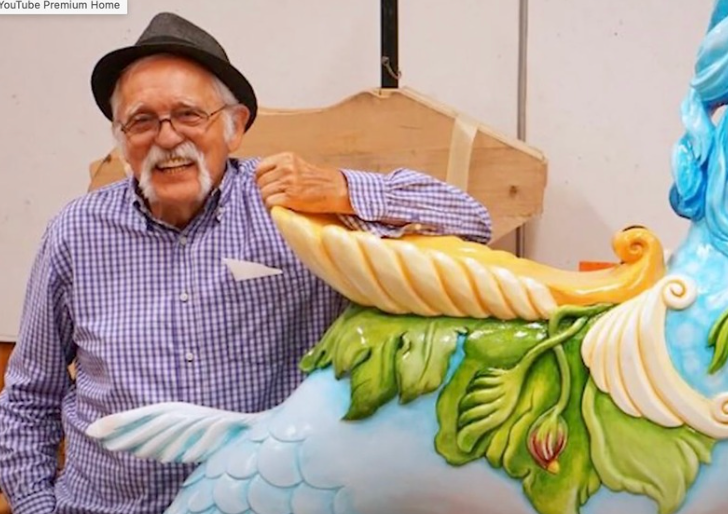 Man in hat smiles, leans on a carousel sculpture with blue and green paint.