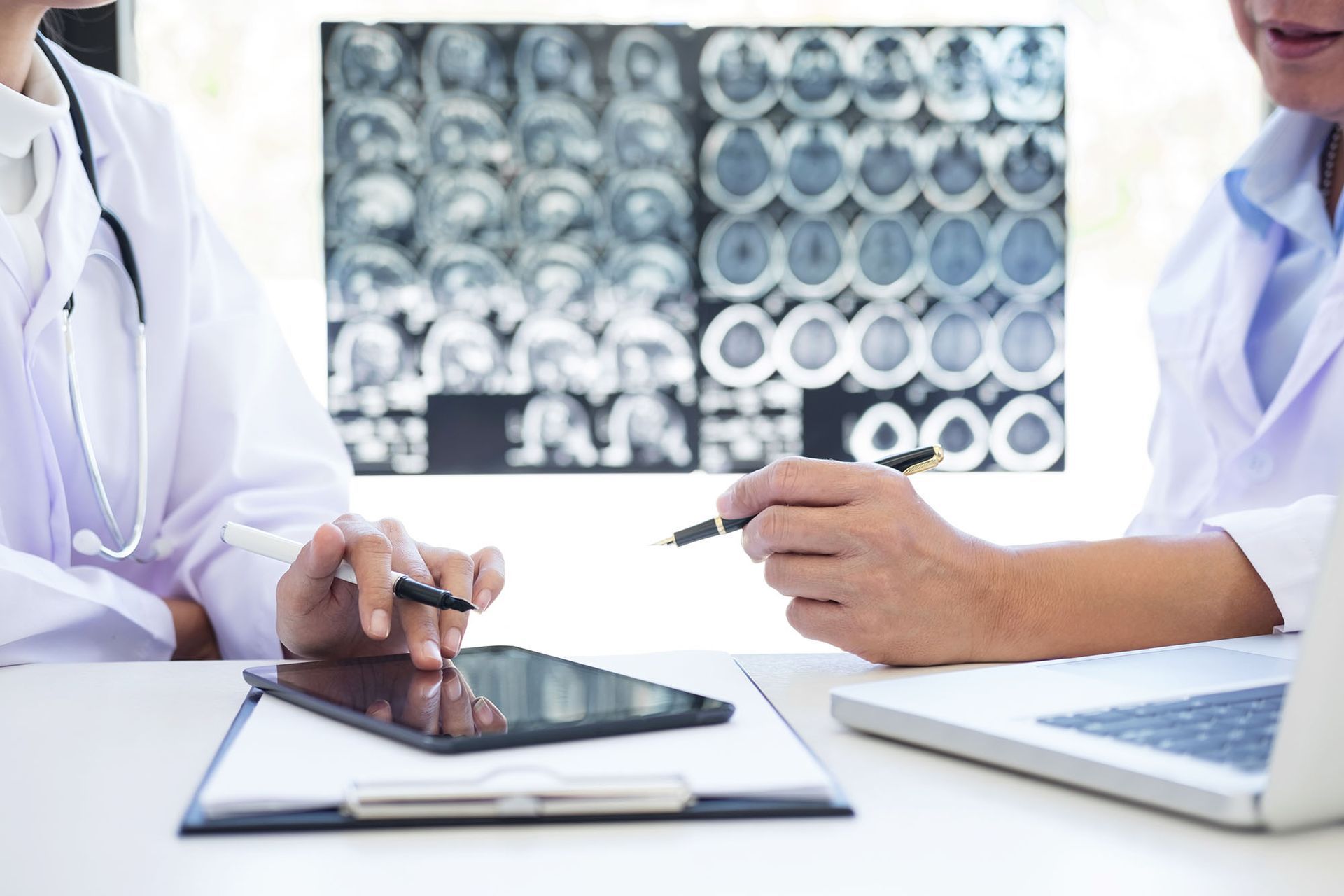 A doctor discusses with another doctor using a tablet the results of a brain x-ray in the background