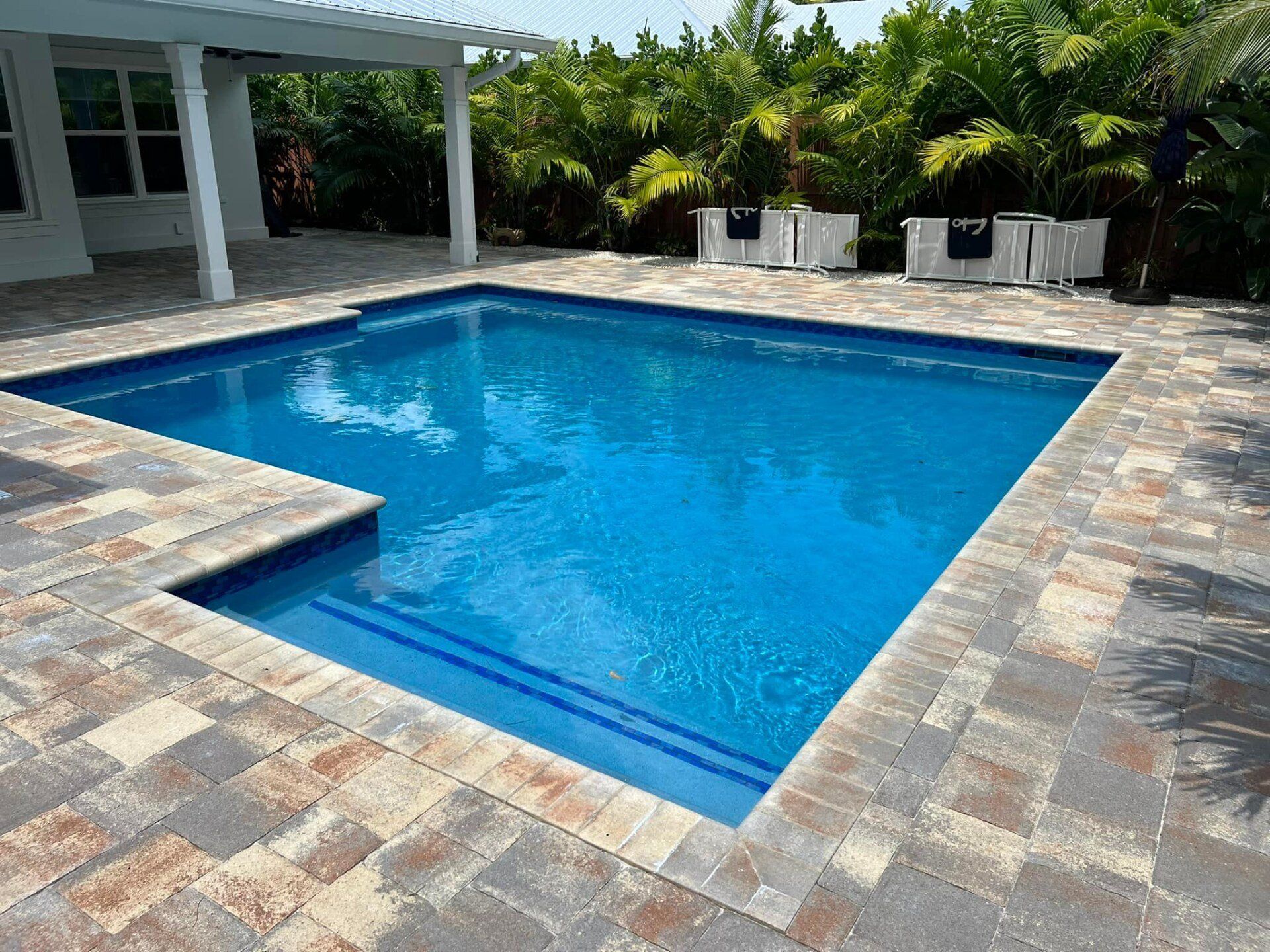 A rectangular swimming pool with blue water, surrounded by brick pavers. A covered patio is visible.