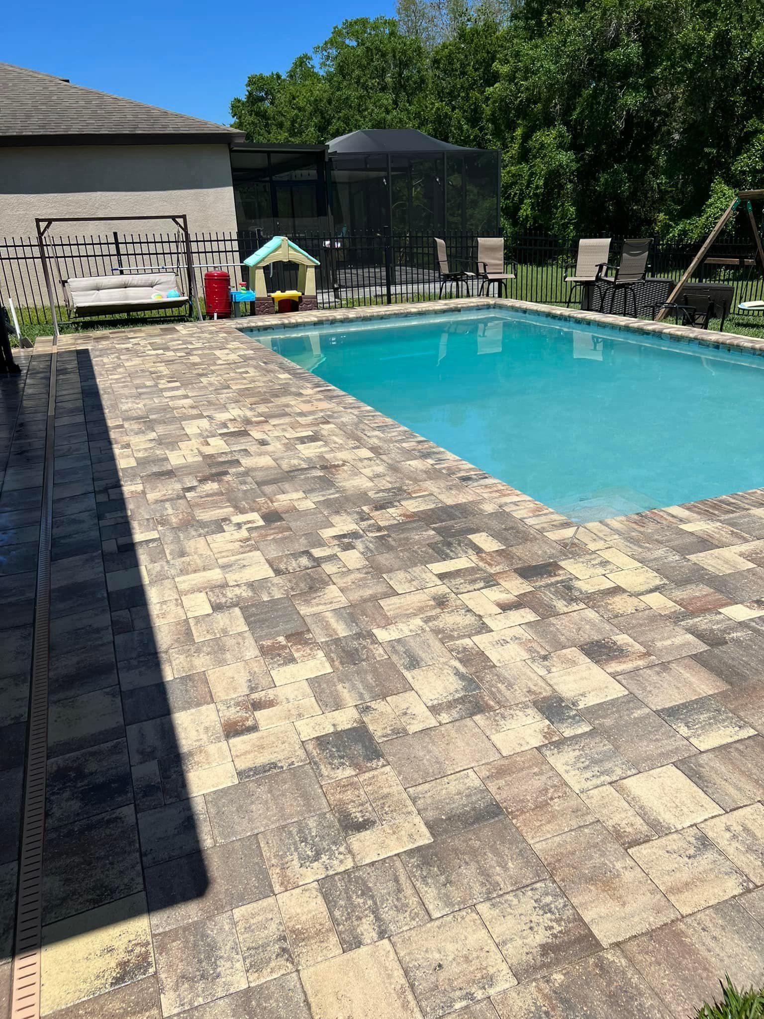 Pool with brick patio, beige and gray. Fenced backyard. Gazebo, chairs. Sunny day.