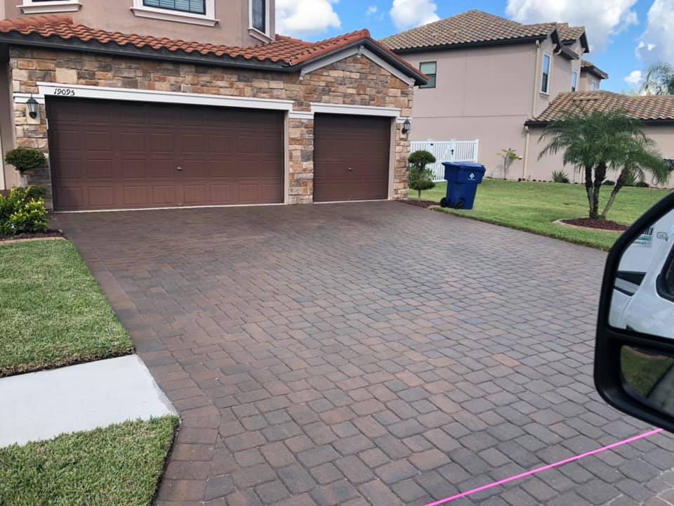 A brick driveway leading to a house with brown garage doors. Green lawn and blue trash bin visible.