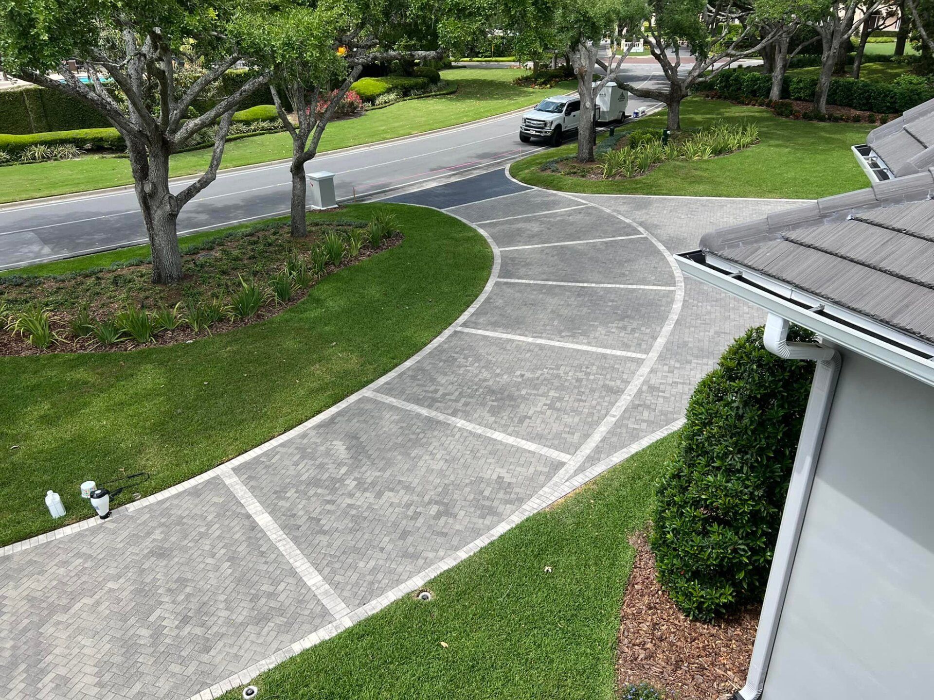 Driveway curving towards a street, bordered by grass and trees. A white car is visible.
