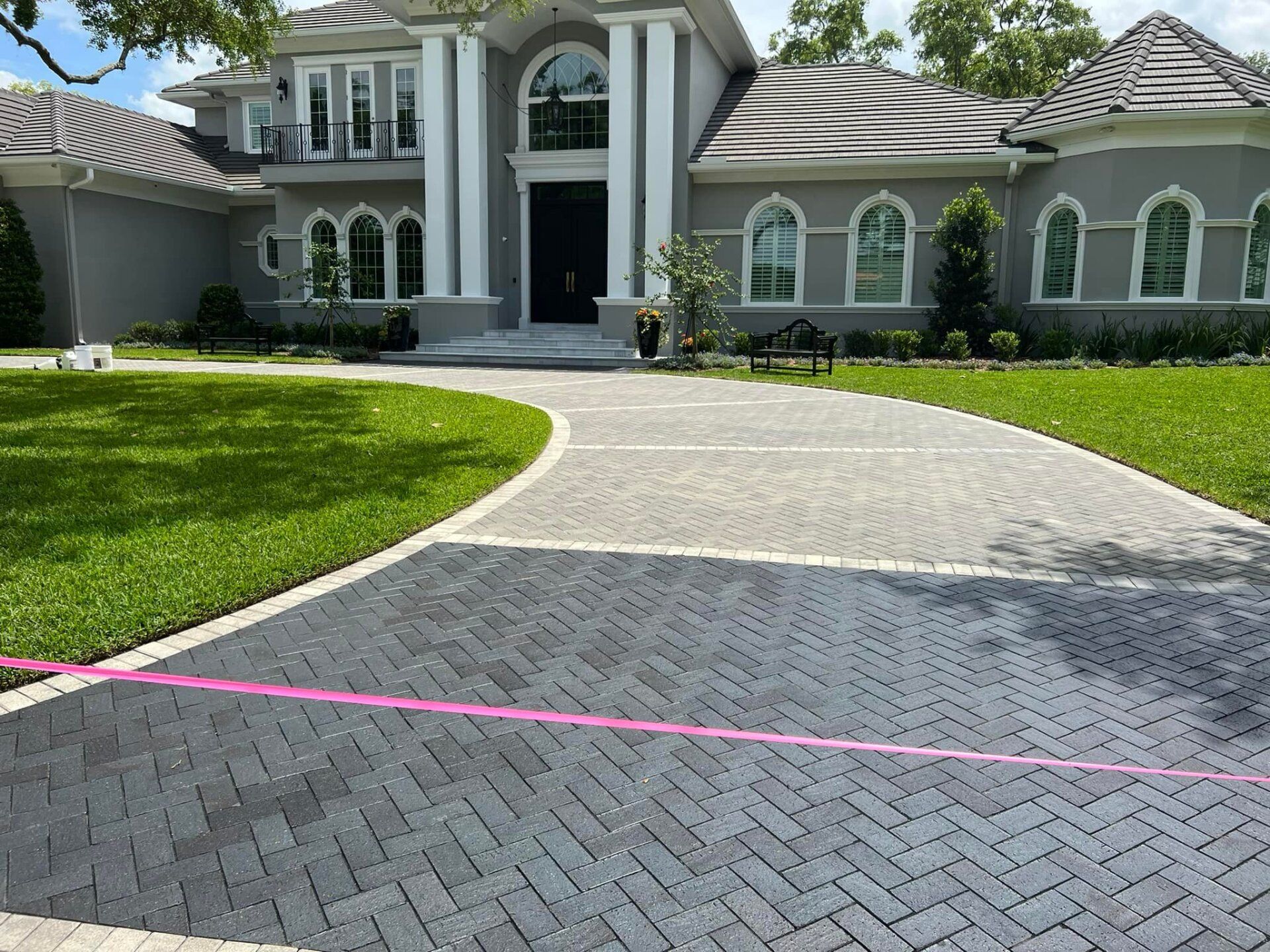 Brick driveway leading to a large house with columns. Pink ribbon across the darker bricks.