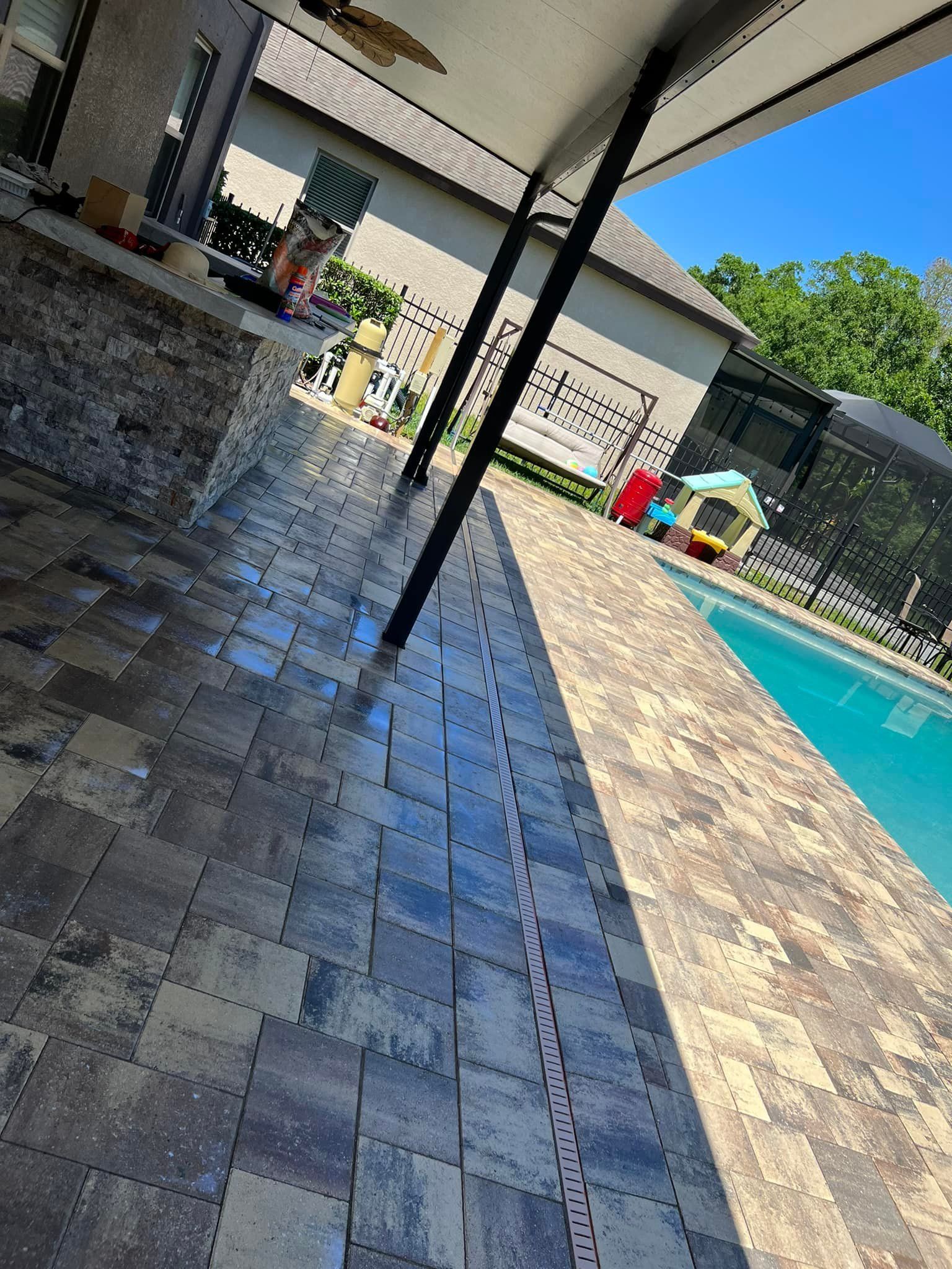 Patio with dark gray pavers, pool, and house in background. Shadow of beams on pavers.