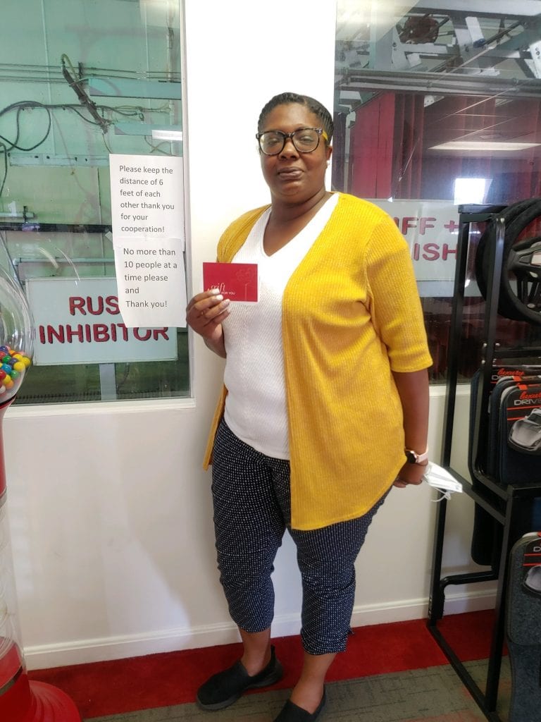 Woman holding a red card, wearing glasses, mustard cardigan, and patterned pants in a car wash.