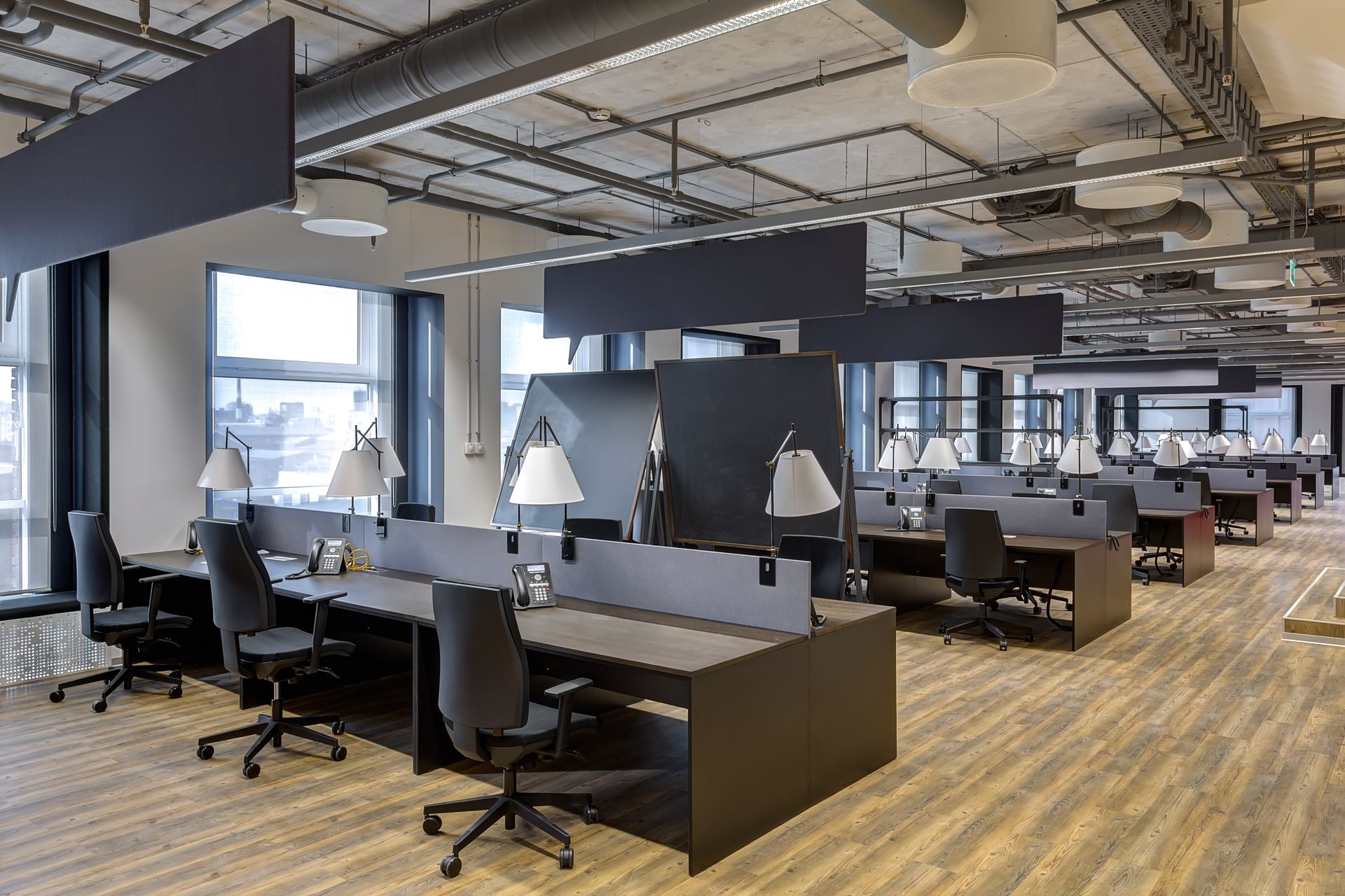 Open-plan office with rows of desks, chairs, and monitors under exposed ceilings and wood flooring