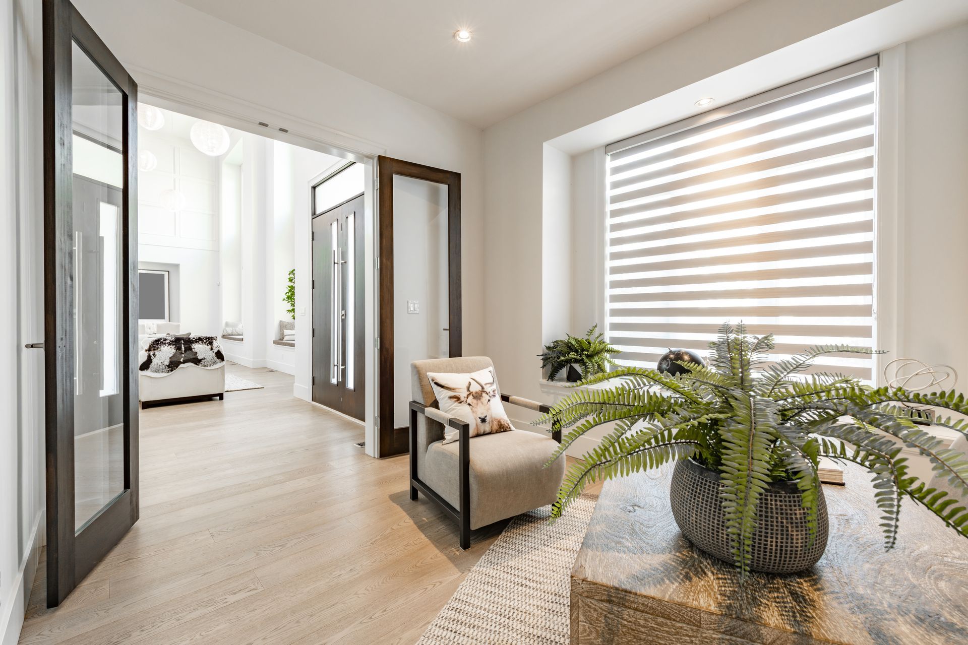 Bright hallway with wood floors, armchair, potted fern, and large window with blinds