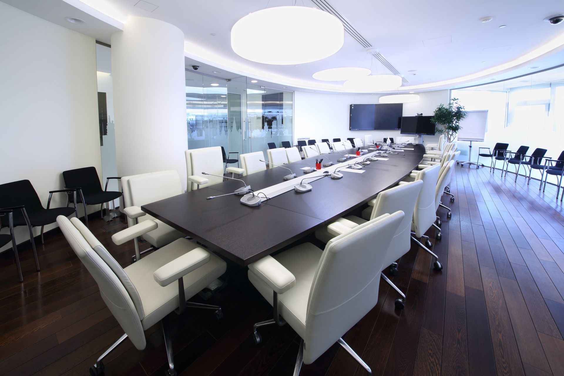 Modern conference room with a long table, white chairs, large windows, and a wall-mounted screen