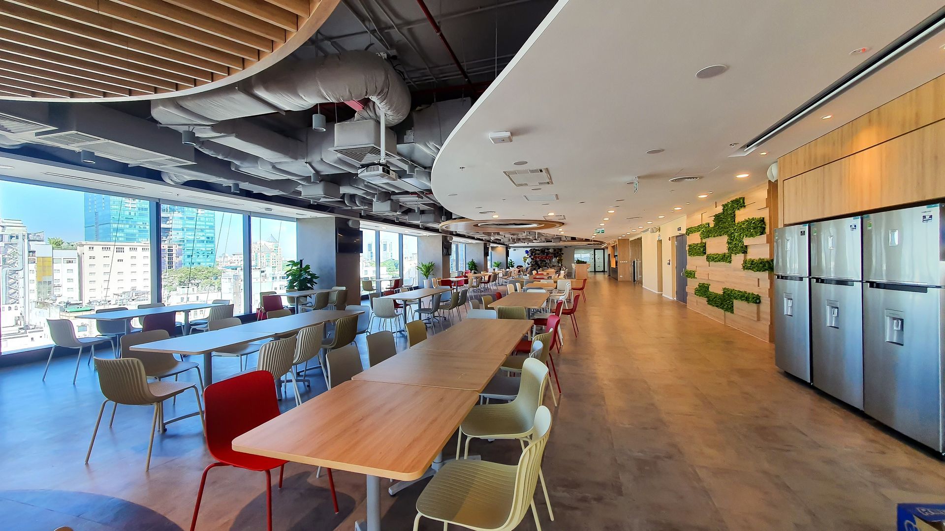 Modern office cafeteria with long wooden tables, chairs, windows, and a row of silver refrigerators