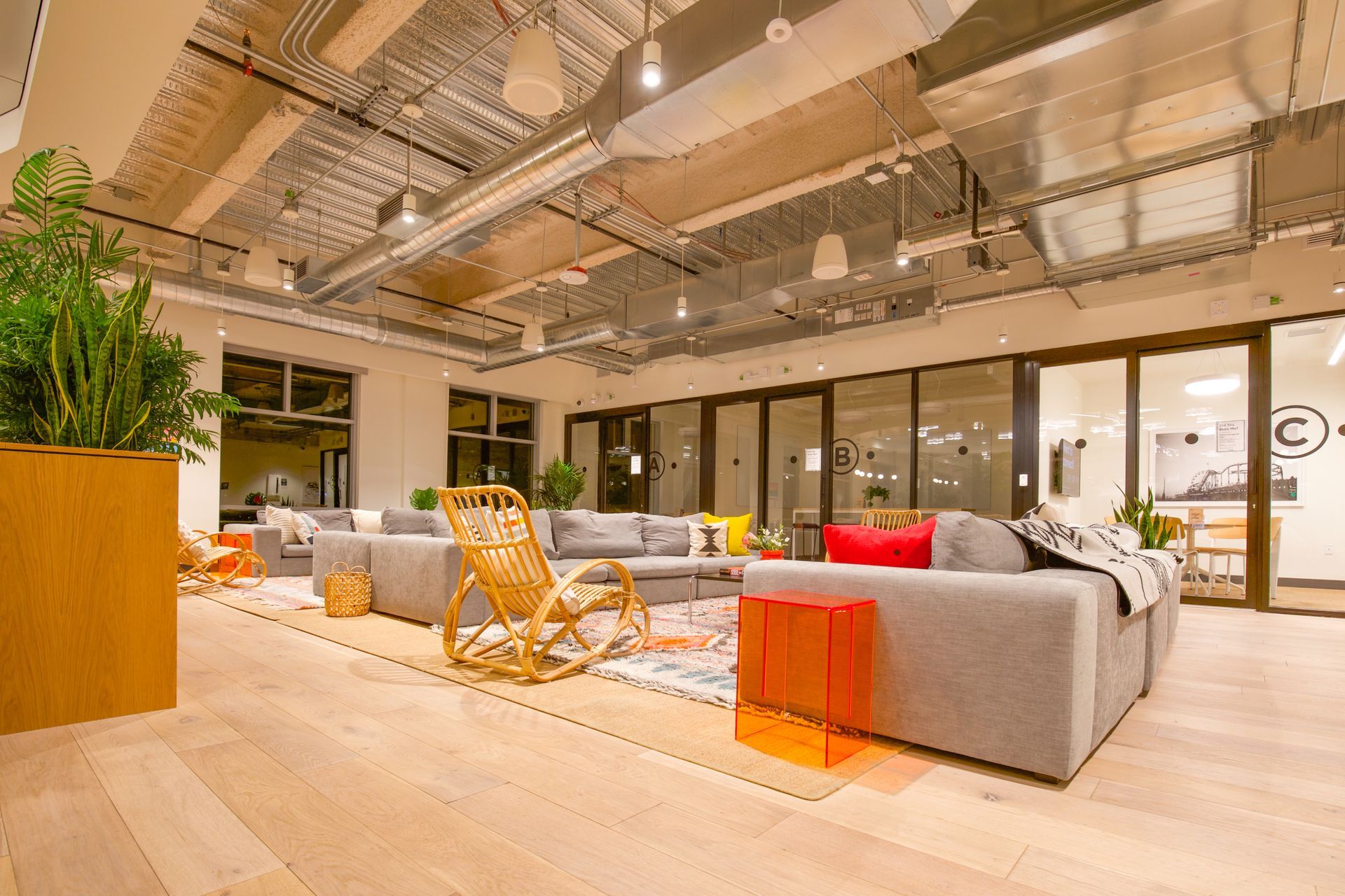 Modern open office lounge with gray sofas, orange chair, wooden floor, and ceiling lights