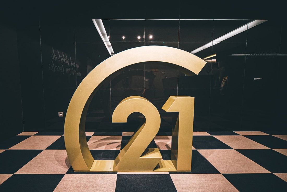 Gold “21” sculpture on a black-and-white checkered floor in a dark gallery space
