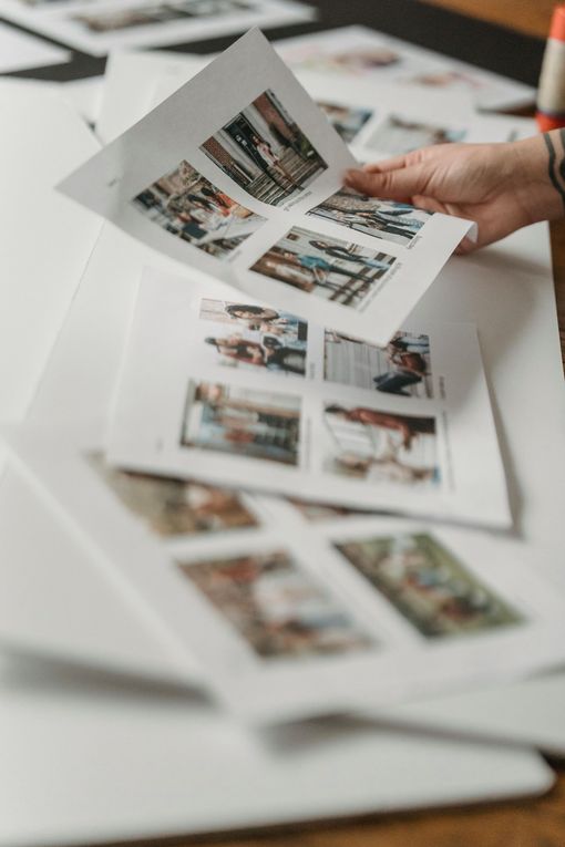 A hand holds a sheet of paper with small photo previews, laid over other similar printed contact sheets on a table.