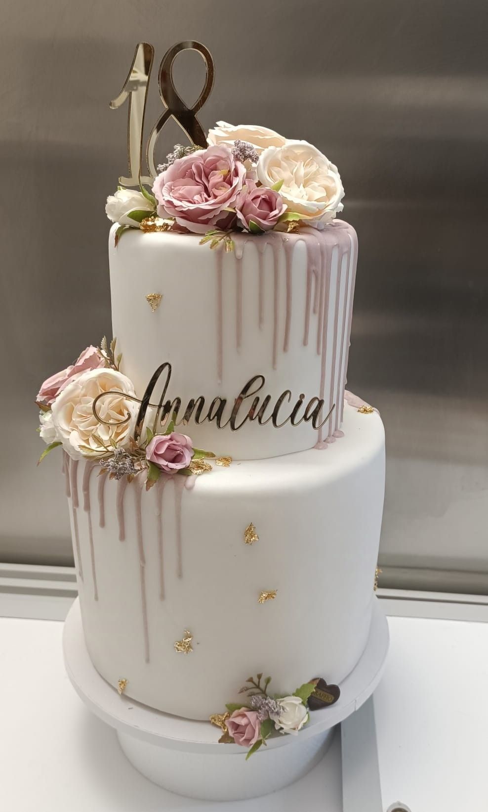 Torta bianca a due piani con glassa rosa, fiori e un topper con la scritta