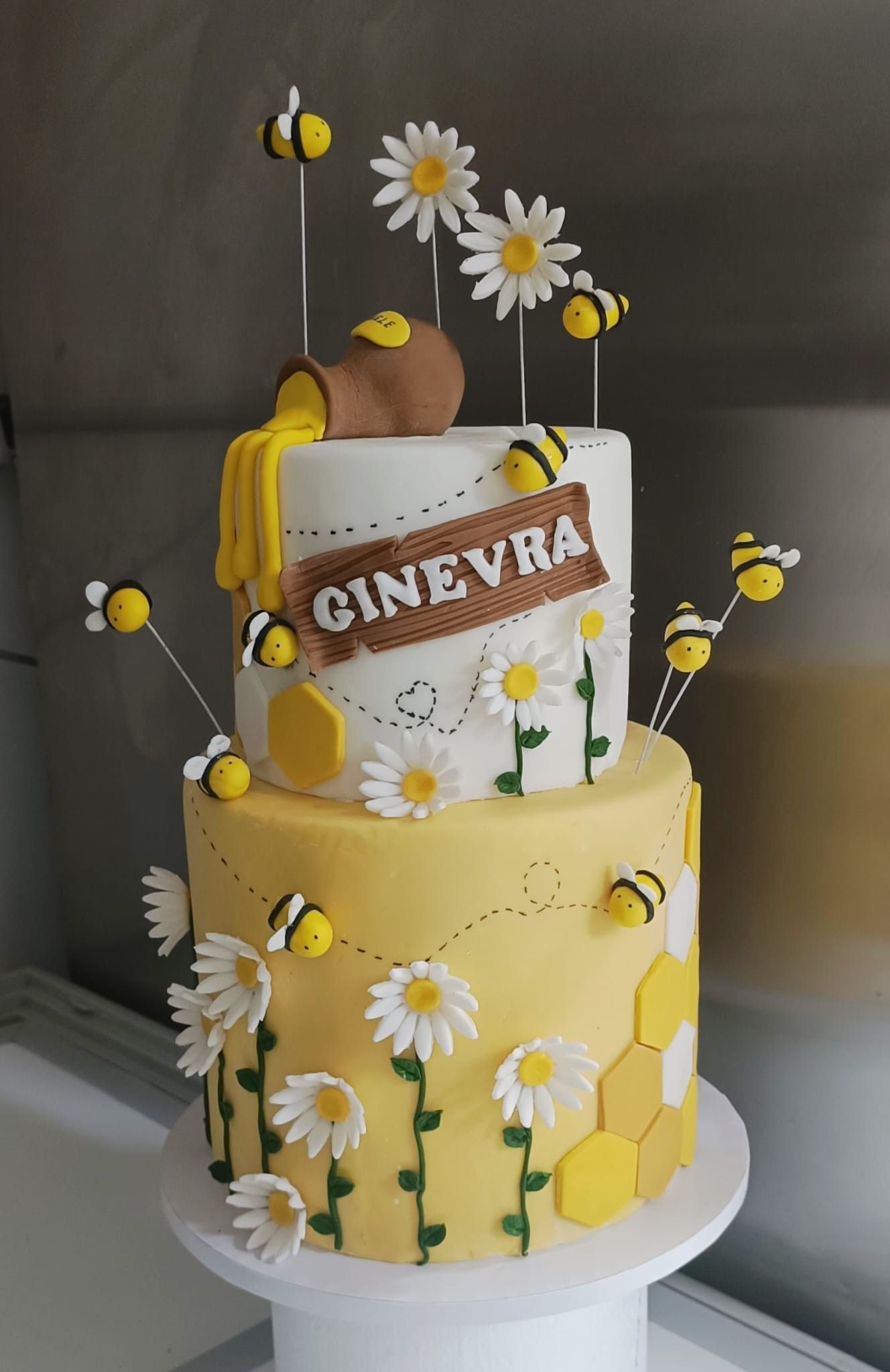 Torta a due piani a tema ape con vasetto di miele, margherite e api in volo; glassa bianca e gialla.