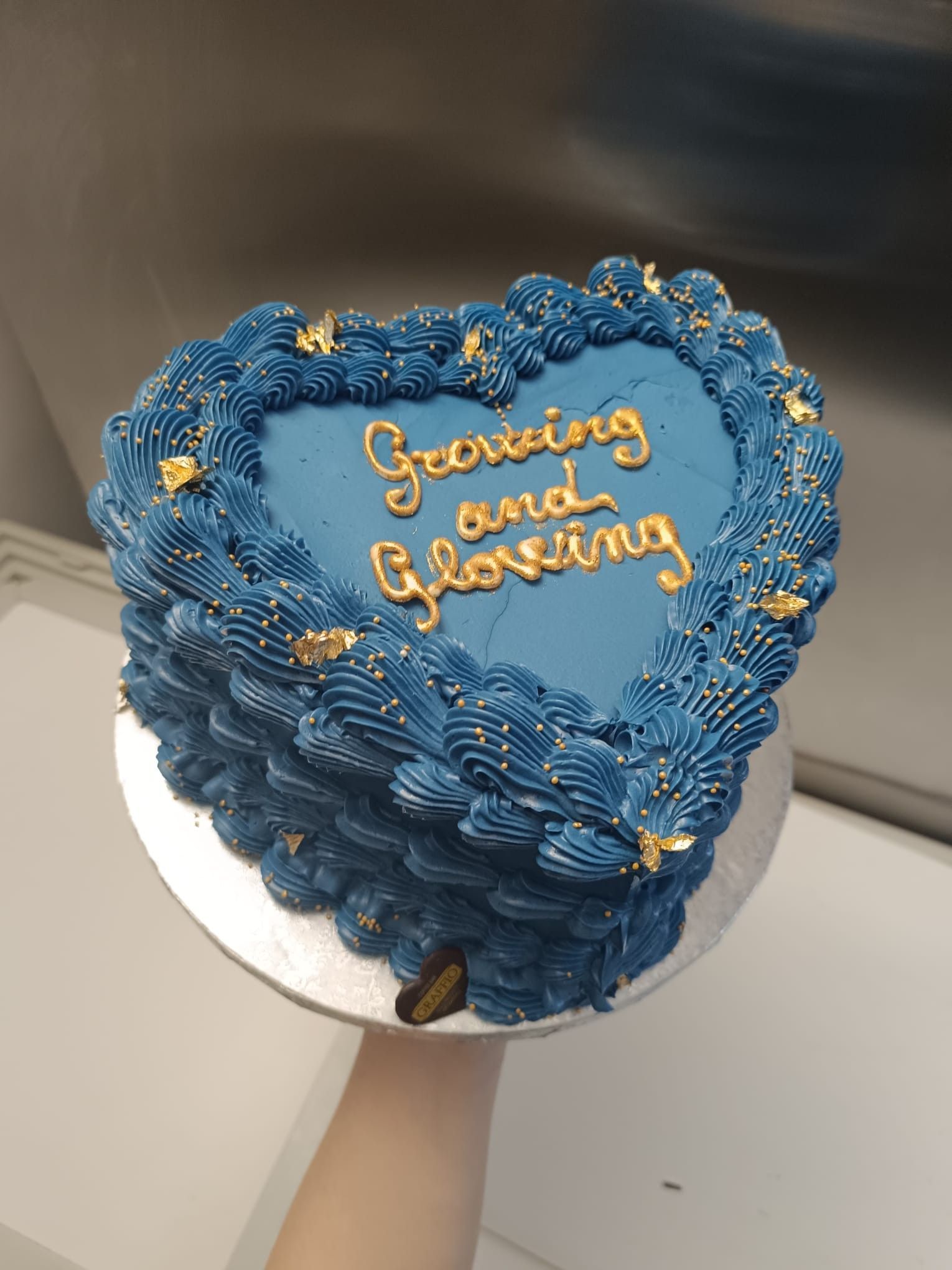 Torta a forma di cuore, glassata di blu, con scritte dorate e granelli di zucchero, tenuta in mano. Sulla scritta c'è scritto