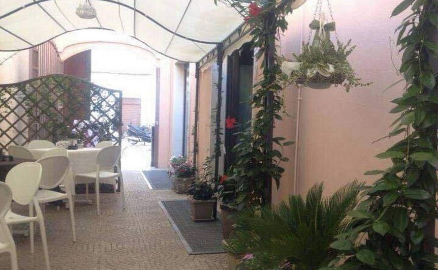 Zona pranzo all'aperto con sedie e tavoli bianchi sotto una tettoia. Rampicanti e piante in vaso decorano lo spazio.