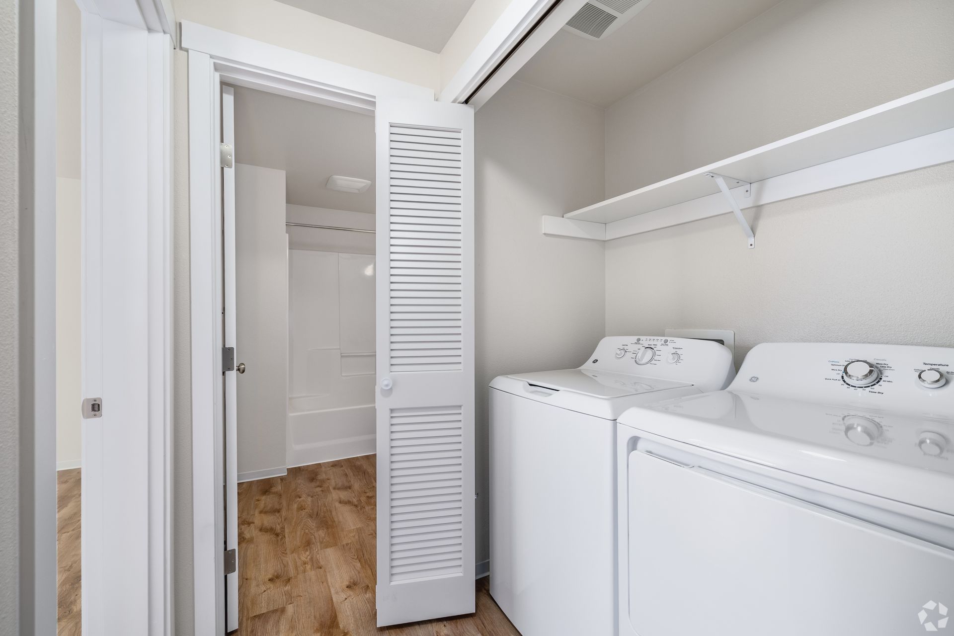 Laundry closet with a washer and dryer beside an open louvered door and white shelving.