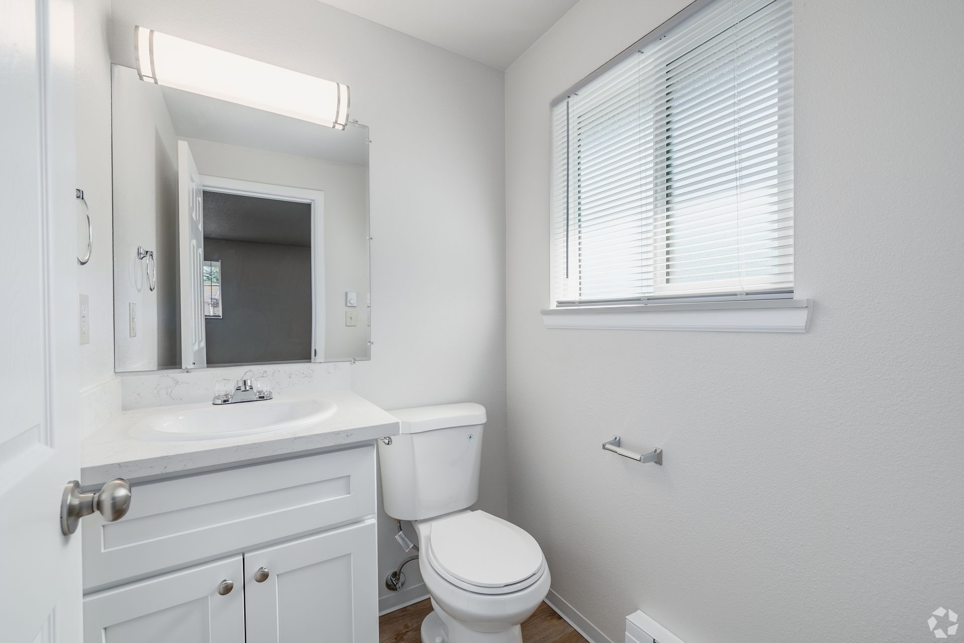 Small white bathroom with vanity, mirror, toilet, and window with blinds.